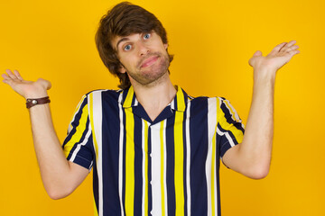 Puzzled and clueless Young handsome Caucasian man, wearing stripped shirt standing against yellow wall with arms out, shrugging shoulders, saying: who cares, so what, I don't know.