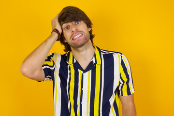 Oops, what did I do? Young handsome Caucasian man, wearing stripped shirt standing against yellow wall holding hand on forehead with frightened and regret expression.