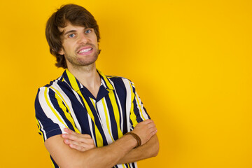 Image of cheerful Young handsome Caucasian man, wearing stripped shirt standing against yellow wall with arms crossed. Looking and smiling at the camera. Confidence concept.