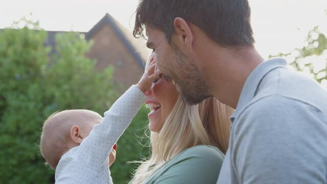Loving parents cuddling baby daughter as they hold her outdoors in garden against flaring sun - shot in slow motion
