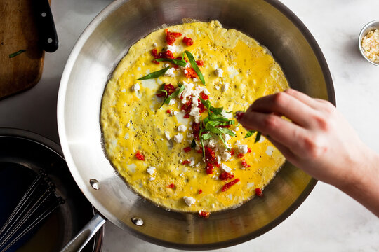 Overhead view of woman spreading basil leaves on egg omelet cooking in pan