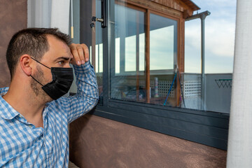 Sick man from coronavirus looking out the window and wearing mask for protection and recovery from illness at home