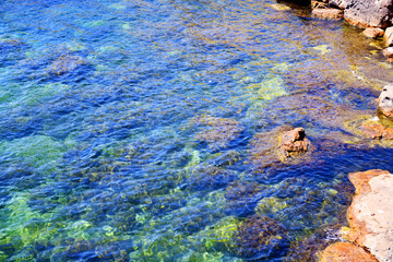 A beautiful shallow lagoon with colorful underwater vegetation visible through the clear water. The rocky coast is orange-red. Mediterranean Sea. View from above.