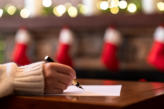 Hand of young woman with fountain pen and sheet of paper on background of christmas fireplace with decoration of light bulbs. Concept of writing wish list.