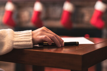 Hand of young woman with fountain pen and sheet of paper on background of christmas tree and fireplace with decoration of light bulbs. Concept of writing letter to Santa Claus
