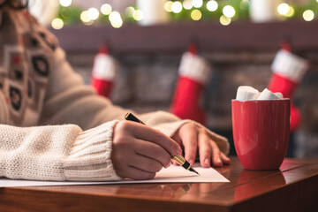 Woman writing wish list using fountain pen on sheet of paper at christmas fireplace with decoration...