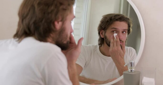 Positive young man with beard using cotton pad and cleansing lotion for face after morning shower. Men's health and beauty procedures.