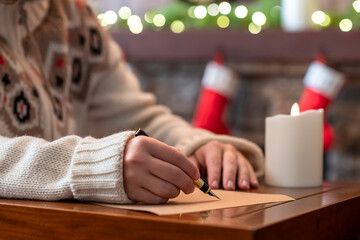 Woman writing wish list using fountain pen on sheet of paper at christmas fireplace with decoration of light bulbs and candle on table