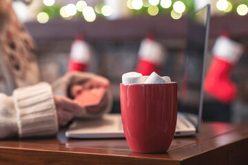 Naklejka na ściany i meble Woman holding credit card using laptop for making order sitting at table with cup of hot cocoa and marshmallow at christmas fireplace with decoration of light bulbs. Close up.