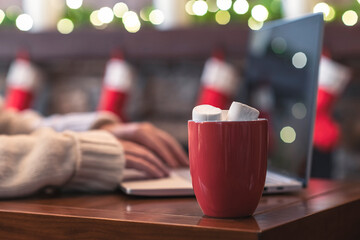 Lonely woman freelancer enjoying with cup of hot cocoa and marshmallow working on laptop near christmas fireplace with decoration of light bulbs.