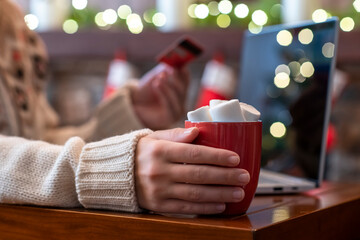 Woman  holding credit card using laptop for making order sitting at table with cup of hot cocoa and marshmallow at christmas fireplace with decoration of light bulbs. Close up.