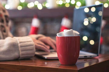 Lonely woman freelancer enjoying with cup of hot cocoa and marshmallow working on laptop near christmas fireplace with decoration of light bulbs. Selective focus