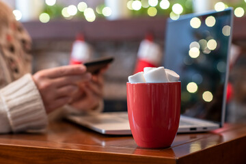 Woman  holding credit card using laptop for making order sitting at table with cup of hot cocoa and marshmallow at christmas fireplace with decoration of light bulbs. Close up.