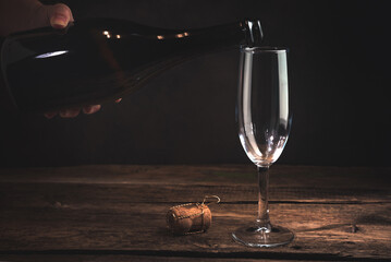 Hand holding a bottle of champagne over an empty glass on a dark background. Concept of festive alcoholic backgrounds. Side view.