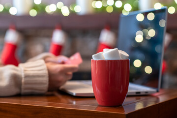 Woman  holding credit card using laptop for making order sitting at table with cup of hot cocoa and marshmallow at christmas fireplace with decoration of light bulbs. Close up.