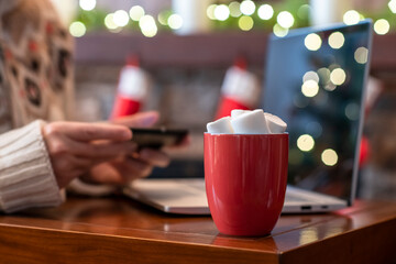 Woman  holding mobile phone using laptop for making order sitting at table with cup of hot cocoa and marshmallow at christmas fireplace with decoration of light bulbs. Close up.