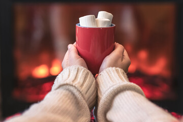 Woman with cup of hot cocoa and marshmallow warming legs in winter white socks near fireplace flame, covered christmas plaid