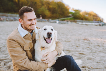 Handsome smiling young man sit on beach and hug his beautiful golden retriever. Trust, care pet...