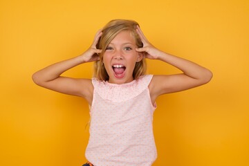 Excited  overjoyed Beautiful Caucasian young girl standing against yellow background screams after receiving good news, keeps hands on head, can't believe it.