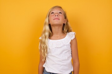 Beautiful Caucasian young girl standing against yellow background looking up as he sees something strange.