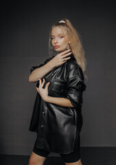 Fototapeta premium Amazing girl with blond hair dressed in a stylish top, shorts and leather shirt posing on a dark background.
