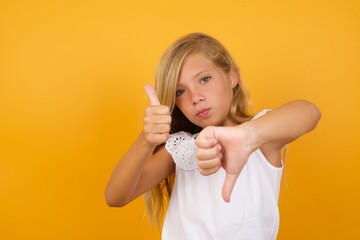 Beautiful Caucasian young girl standing against yellow background feeling unsure making good bad sign. Displeased and unimpressed.