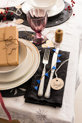 Christmas silverware on a black elegant napkin next to a set of three dishes with a gift box inside and a pretty transparent purple glass. The name Sarah is on a round tag.