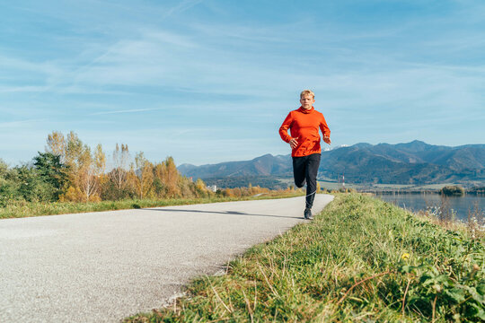 Man dressed in red long sleeve shirt running by the asphalt road with mountain background. Sporty people activities and a healthy lifestyle concept image.