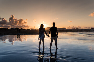 Fototapeta premium Couple from behind holding hands on a beach at sunset.