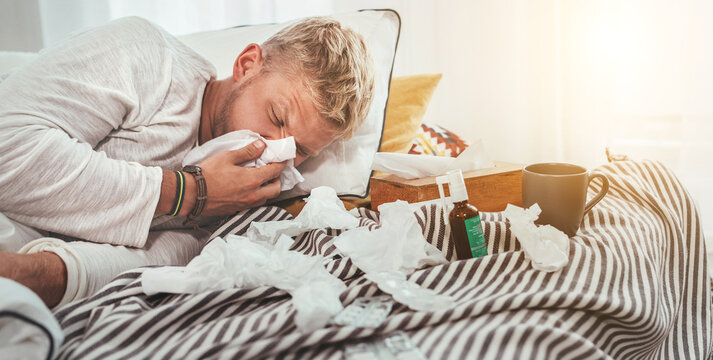 Unhealthy and tired man sneezing or wiping snotty nose man lying in a cozy home bed beside a lot of used paper tissues and medicines on the blanket. Season virus flu and home quarantine concept image.