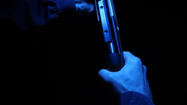 In the dark with blue illumination, a gun is in the hands of a man dressed in a black, ready to fire. Different angles and distinctive details of this weapon are shown. Closeup