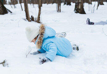 Little girl is making snowman from snowball in snowy winter park. Walking, having fun.Stylish jacket with fur,pants with snowflakes,mittens.Family picnic in cold weather.Outdoor activity