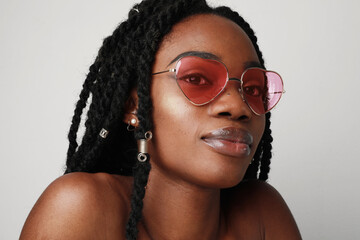 Young black woman with braids wearing pink sunglasses, posing on white wall.