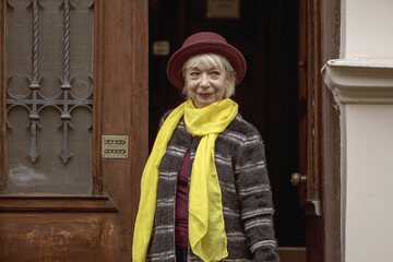 An elderly woman 60-65 years old in a hat comes out of the entrance of an old house. Concept, retirement, fashionista of advanced years, excellent health, meeting on the street, isolation and communic