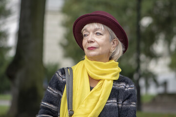 Street portrait of an elderly woman in a fashionable hat and scarf sitting on a bench in the Park and resting, close-up, selective focus. Concept: excellent health in retirement, tourism and travel.