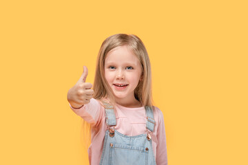 Cute girl looks at the camera and shows a super gesture, finger in. Yellow isolated background.