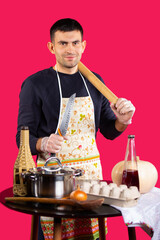 A young man prepares food at home in the kitchen with a knife and rolling pin. Photo on a pink...