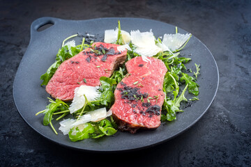Modern style traditional fried New York strip steak with rucola and parmesan offered as close-up on a design plate