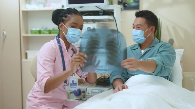 Multi-racial couple of asian patient and afro-american female doctor examining x-ray checkup of lungs for further treatment diagnostics healthcare. Pandemic. Face masks. Coronavirus.
