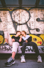 Two girls sitting on a bench against a wall full of graffiti on the street.