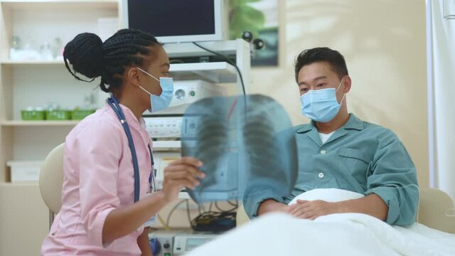 Afro-american femnale therapist explaining disease symptoms to asian chinese patient on x-ray scan image of lungs discussing recovery in hospital office. Medical concept. Multi-ethnic couple.