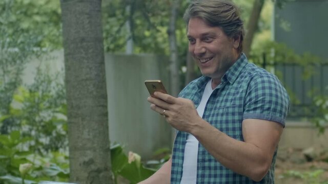 Spraying water on vegetables with a garden hose. A happy man watering the garden from a hose watches funny videos on his phone as he tends to the garden. Concept: gardening and garden care.