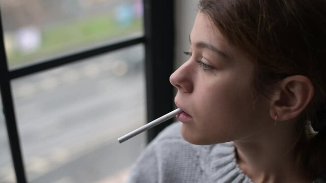 A young woman takes a cigarette in her mouth against the background of passing cars outside the window. Woman about to smoke, looking at cloudy view in window