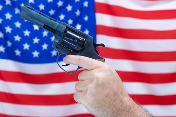 A hand holding a gun in front of a waving flag of the United States of America.