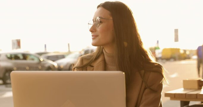 Charming young lady with brown hait in eyeglasses sitting outdoors and working on wireless laptop. Concept of lifestyle, remote work and technology.