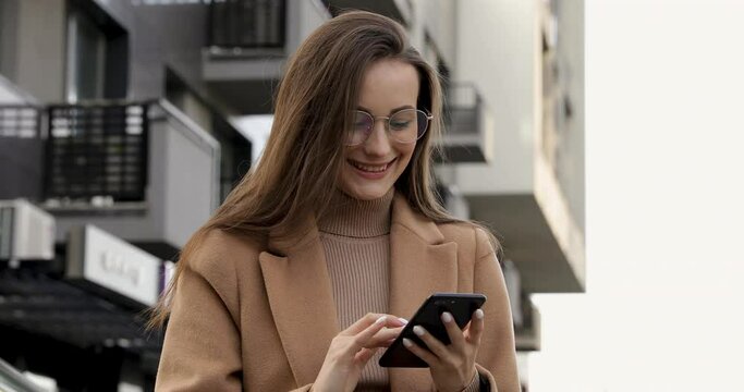 Portrait of smiling young female in eyeglasses staning near dark modern building and using personal mobile phone. Happy woman in autumn coat scrolling with finger on smartphone screen outdoors.