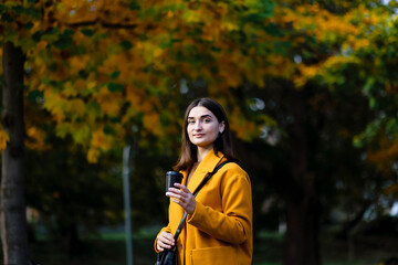 Fototapeta premium Female hand holding a cup of blank paper on a background of the park. Free space. Hands of a young girl in a yellow coat holding a paper cup of coffee. Girl hand hold coffee plastic cup. Coffee.