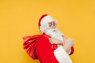 Fototapeta na wymiar Positive Santa with a bag stands on a yellow background, shows a thumb up and looks at the camera with a smile on his face. Christmas concept.