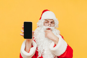 Santa Claus stands on a yellow background, looks at the camera with a pensive face and shows a smartphone with a black screen. Copy space