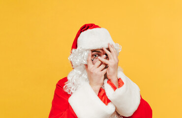 Portrait of a scared Santa Claus on a yellow background, looking at the camera covering his face with his hands. Isolated.